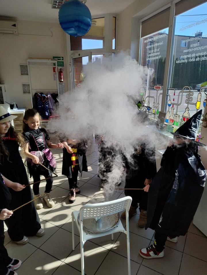 Dym naukowy w eksperymencie chemicznym w szkołach, dzieci w czapkach czarownic, doświadczenie edukacyjne w laboratorium, świetna zabawa dla dzieci, nauka chemii dla dzieci, klimat nauki i eksperymentów, bezpieczeństwo w naukowych eksperymentach, fascynujące doświadczenia z dymem, rozwijanie ciekawości naukowej młodzieży, kreatywne zajęcia edukacyjne dla dzieci, eksploracja naukowa w szkole, pokaz chemicznych reakcji, naukowe eksperymenty dla dzieci, atrakcyjne zajęcia edukacyjne w laboratorium, edukacja w zakresie nauk przyrodniczych.